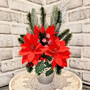 May include: A Christmas floral arrangement with bright red poinsettias, green fir, and silver glitter detailing. The flowers are in a silver pot, with red berries and holly. A festive display for the holiday season, with a classic look.