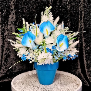 May include: A floral arrangement in a blue pot featuring three prominent blue and white calla lilies with yellow centres, surrounded by white blossoms, small blue flowers, green foliage, and white feathery plumes, creating a dense bouquet.