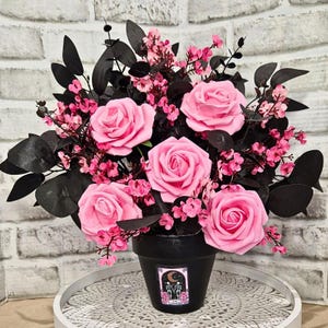 May include: A floral arrangement with large pink roses, small pink flowers, and black leaves in a black pot. The pot has a decorative label with a crescent moon and the words "The Lovers". The arrangement is on a white tray.