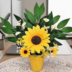 May include: A bright yellow sunflower arrangement in a small yellow pot. The arrangement features large yellow sunflowers, small yellow sunflowers, white daisies, and green leaves. The pot is on a decorative tray.