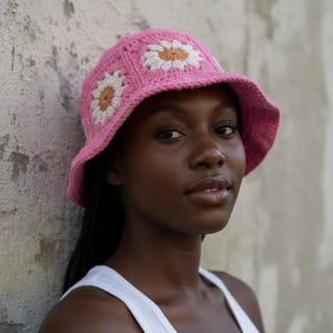 Pode incluir: Um chapéu de balde rosa de crochê com aplicações de margaridas brancas e bege. O chapéu é feito à mão e apresenta uma aba larga. Um acessório elegante para os dias ensolarados.