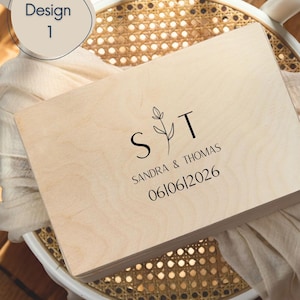 May include: A rectangular light-coloured wooden box featuring a black printed design with the letters S and T separated by a botanical line drawing. Below the letters, the text reads SANDRA & THOMAS followed by the date 06|06|2026.