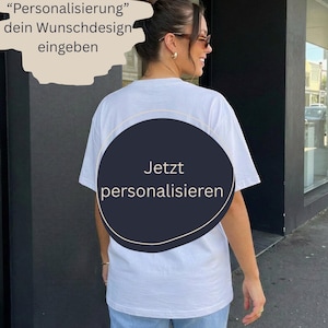 Könnte beinhalten: Ein weißes kurzärmeliges T-Shirt von hinten gesehen, mit einer großen dunklen kreisförmigen Grafik in der Mitte und dem Text "Jetzt personalisieren". Ein beiges Textfeld darüber enthält "im Feld "Personalisierung" dein Wunschdesign eingeben".