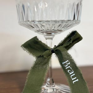 Könnte beinhalten: Ein klares Champagnerglas im Vintage-Stil mit einem dekorativen grünen Band, das um den Stiel gebunden ist. Auf dem Band ist das Wort "Braut" in Weiß aufgedruckt. Das Glas steht auf einer Holzoberfläche.