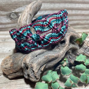 May include: A macrame bracelet featuring an intricate woven pattern of teal and deep burgundy cord. The bracelet is constructed with a series of knotted loops and open spaces, creating a textured, flexible band that wraps around the wrist.