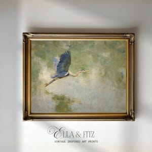 Puede incluir: Una impresión artística enmarcada de inspiración vintage que representa una garza azul en vuelo. La pintura tiene una paleta de colores apagados de verdes, azules y marrones. El marco es ornamentado con un acabado dorado. El texto "Ella & Fitz" y "Vintage Inspired Art Prints" está en la parte inferior.