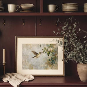 K&ouml;nnte beinhalten: Ein gerahmter Druck eines Kolibris im Flug, mit einem goldenen Rahmen. Das Kunstwerk zeigt den Vogel in der N&auml;he eines bl&uuml;henden Zweigs, vor einem weichen, ged&auml;mpften Hintergrund. Der Druck wird auf einer dunkelroten Oberfl&auml;che mit einer Kerze und einer Vase mit Gr&uuml;nzeug in der N&auml;he ausgestellt.