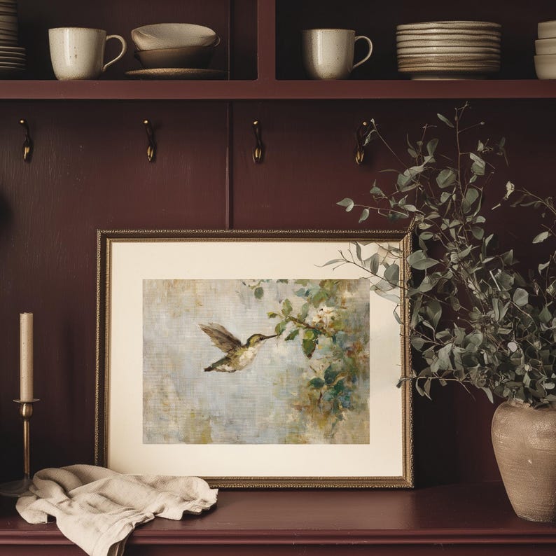K&ouml;nnte beinhalten: Ein gerahmter Druck eines Kolibris im Flug, mit einem goldenen Rahmen. Das Kunstwerk zeigt den Vogel in der N&auml;he eines bl&uuml;henden Zweigs, vor einem weichen, ged&auml;mpften Hintergrund. Der Druck wird auf einer dunkelroten Oberfl&auml;che mit einer Kerze und einer Vase mit Gr&uuml;nzeug in der N&auml;he ausgestellt.