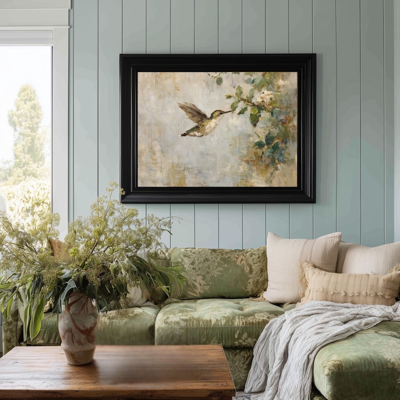 K&ouml;nnte beinhalten: Gerahmtes Kunstwerk mit einem Kolibri im Flug, der sich einer bl&uuml;henden Zweig n&auml;hert. Das Gem&auml;lde hat eine ged&auml;mpfte Farbpalette und einen schwarzen Rahmen. Es h&auml;ngt &uuml;ber einem gr&uuml;nen, gemusterten Sofa mit dekorativen Kissen.