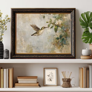 K&ouml;nnte beinhalten: Gerahmtes Gem&auml;lde eines Kolibris im Flug, der nach einem bl&uuml;henden Zweig greift. Das Kunstwerk hat eine ged&auml;mpfte Farbpalette mit einem schwarzen und goldenen, verzierten Rahmen. Das Gem&auml;lde wird auf einem wei&szlig;en Regal mit anderen dekorativen Gegenst&auml;nden ausgestellt.