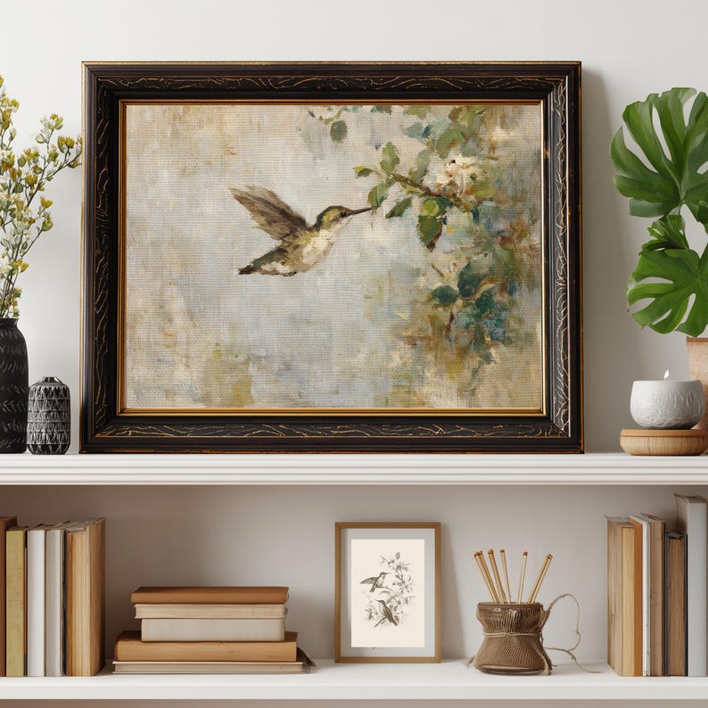 K&ouml;nnte beinhalten: Gerahmtes Gem&auml;lde eines Kolibris im Flug, der nach einem bl&uuml;henden Zweig greift. Das Kunstwerk hat eine ged&auml;mpfte Farbpalette mit einem schwarzen und goldenen, verzierten Rahmen. Das Gem&auml;lde wird auf einem wei&szlig;en Regal mit anderen dekorativen Gegenst&auml;nden ausgestellt.
