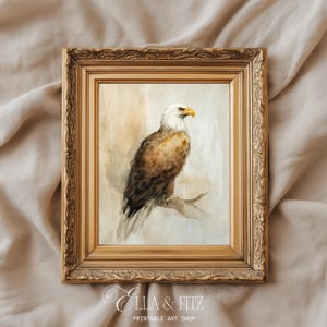 May include: A framed watercolor print of a bald eagle perched on a branch. The eagle has white feathers on its head and a yellow beak. The frame is gold-colored with ornate detailing. The print is from Ella & Fitz Printable Art Shop.