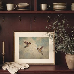 May include: A framed print depicting two hummingbirds in flight against a pale blue and white sky. The artwork is in a gold frame and rests on a dark red surface. A candle and a vase with greenery are also visible.