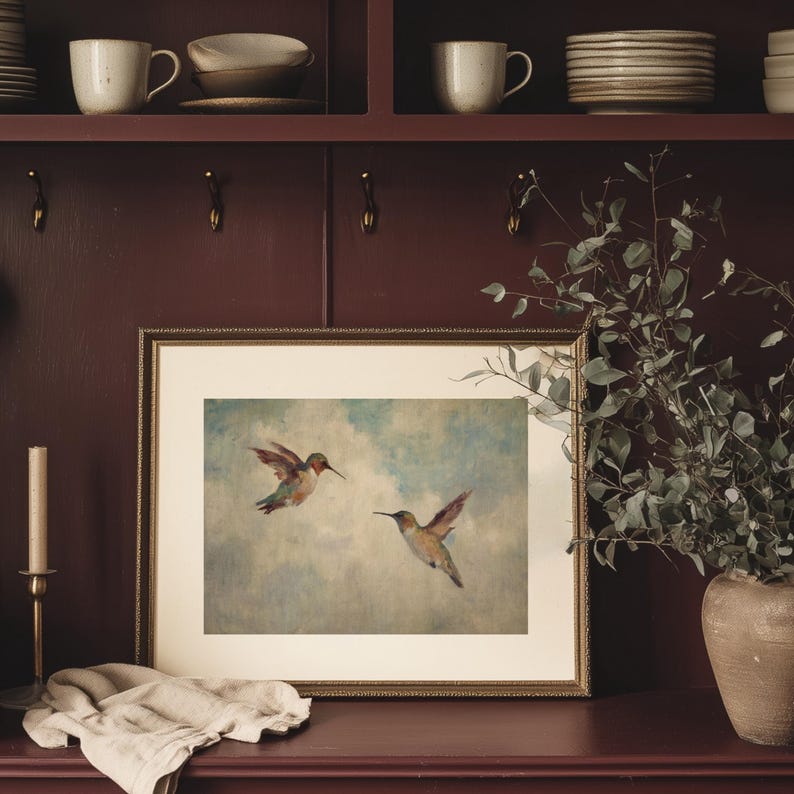May include: A framed print depicting two hummingbirds in flight against a pale blue and white sky. The artwork is in a gold frame and rests on a dark red surface. A candle and a vase with greenery are also visible.