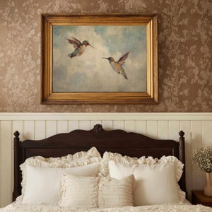 May include: A framed painting of two hummingbirds in flight against a blue sky. The painting has a gold-coloured frame and hangs above a dark brown wooden headboard. White ruffled pillows and bedding are visible below the headboard.