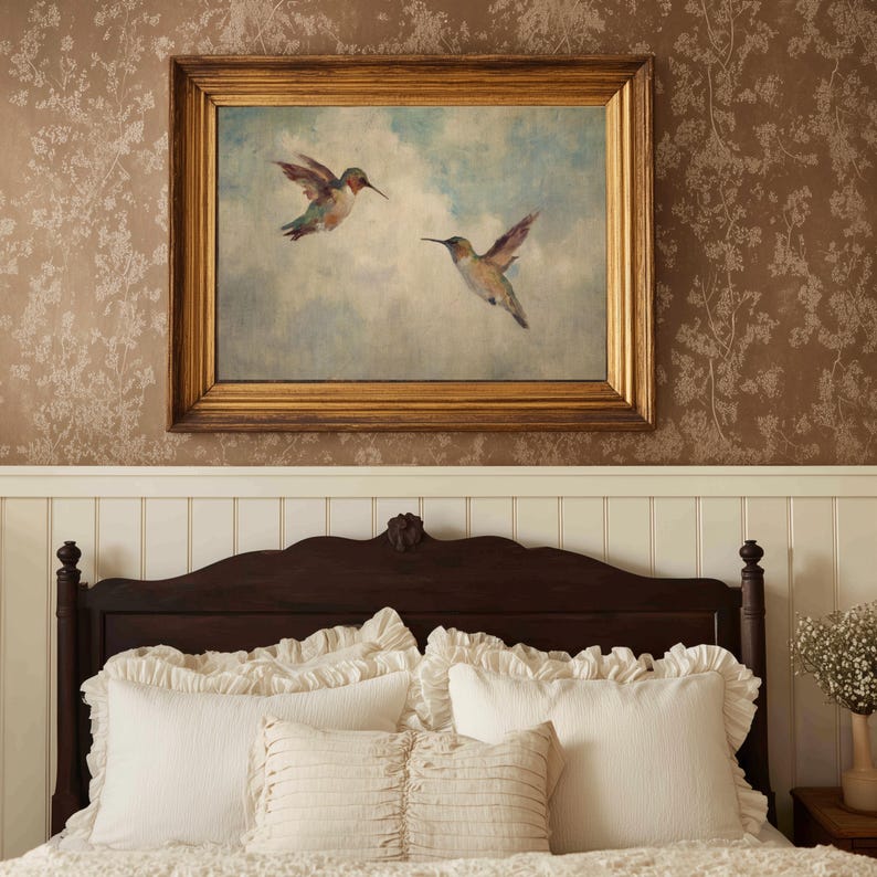 May include: A framed painting of two hummingbirds in flight against a blue sky. The painting has a gold-coloured frame and hangs above a dark brown wooden headboard. White ruffled pillows and bedding are visible below the headboard.