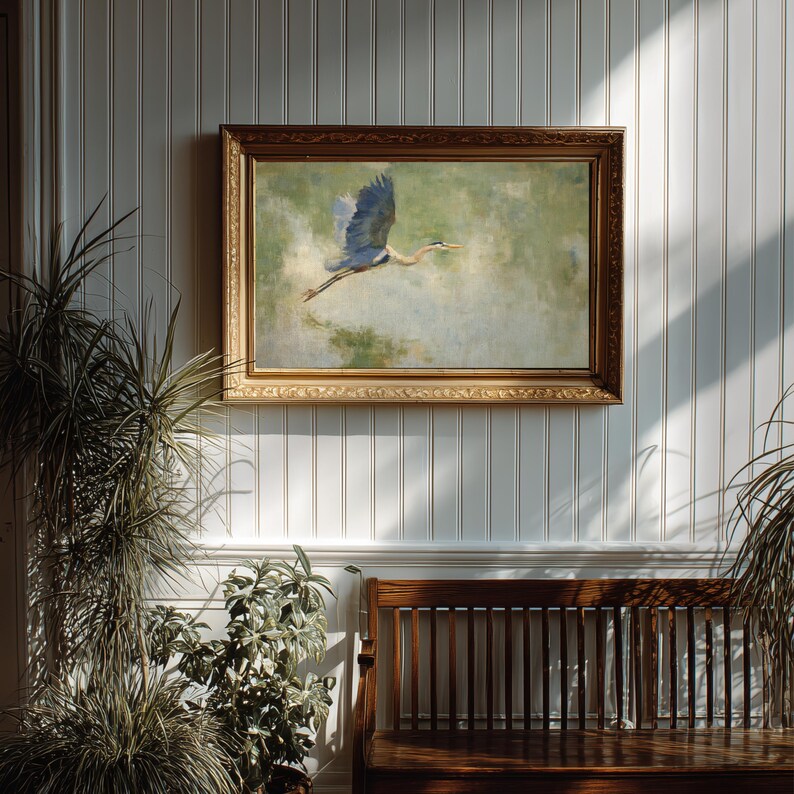 May include: A framed painting of a blue heron in flight, set against a green and blue background. The artwork is displayed on a white paneled wall, above a wooden bench. The frame is a warm, golden brown color.
