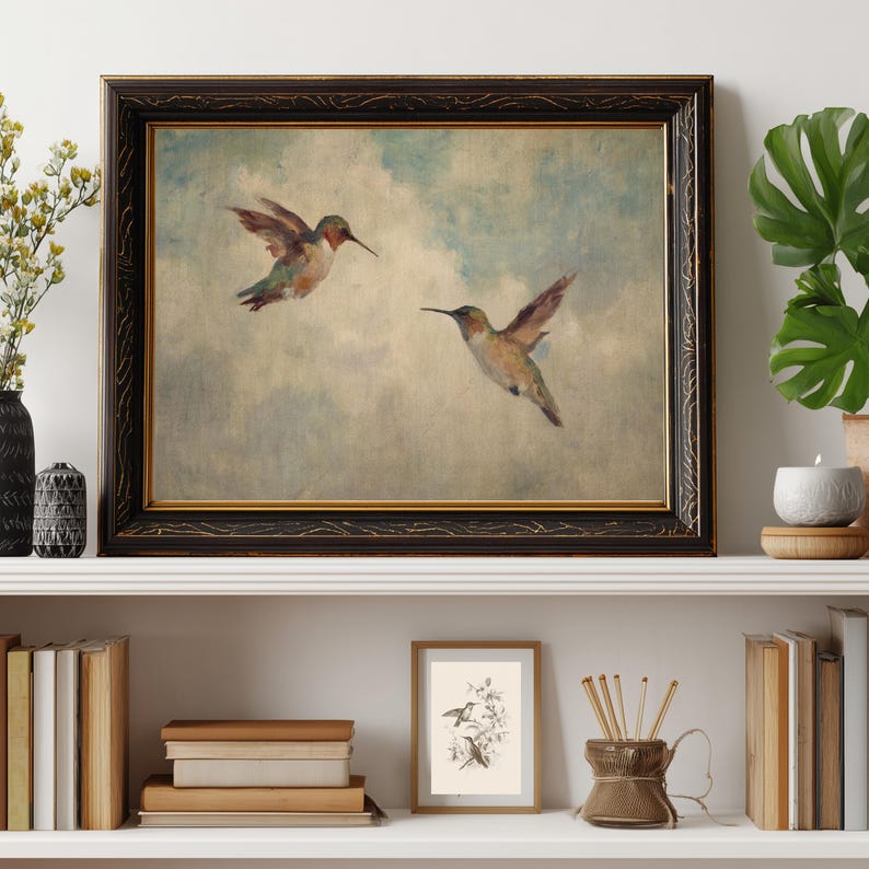 May include: A framed print depicting two hummingbirds in mid-flight against a cloudy backdrop. The artwork has a vintage style, with soft colours and a dark, ornate frame. The print is displayed on a white shelf with other decorative items.
