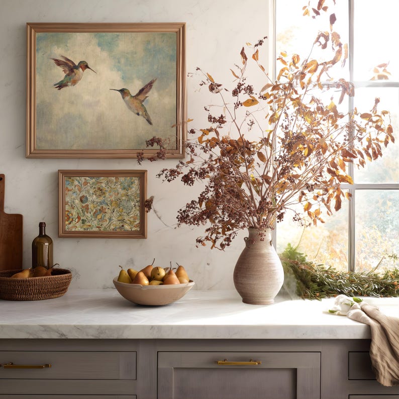 May include: A kitchen interior with a marble work surface, a bowl of pears, and a vase of dried branches. Two framed pictures are on the wall, one showing hummingbirds. The cabinets below are grey with gold handles.