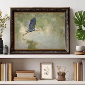 May include: A framed painting of a blue heron in flight against a green and blue background. The artwork is in a dark, ornate frame with gold accents. The painting is displayed on a white shelf with books, a candle, and other decorative items.