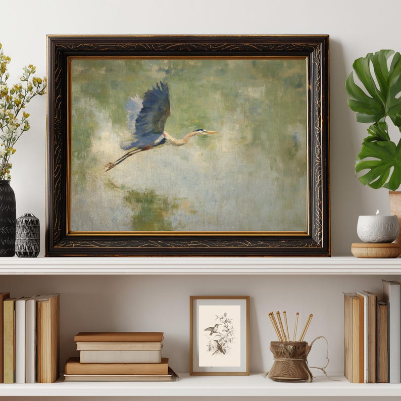 May include: A framed painting of a blue heron in flight against a green and blue background. The artwork is in a dark, ornate frame with gold accents. The painting is displayed on a white shelf with books, a candle, and other decorative items.