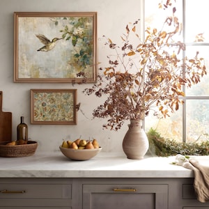 K&ouml;nnte beinhalten: Eine K&uuml;chenszene mit gerahmten Kunstwerken, einer Vase mit getrockneten Zweigen und einer Sch&uuml;ssel mit Birnen. Die Kunstwerke zeigen einen Kolibri und florale Muster. Die Arbeitsplatte ist aus Marmor, und die Schr&auml;nke sind grau mit goldenen Griffen.