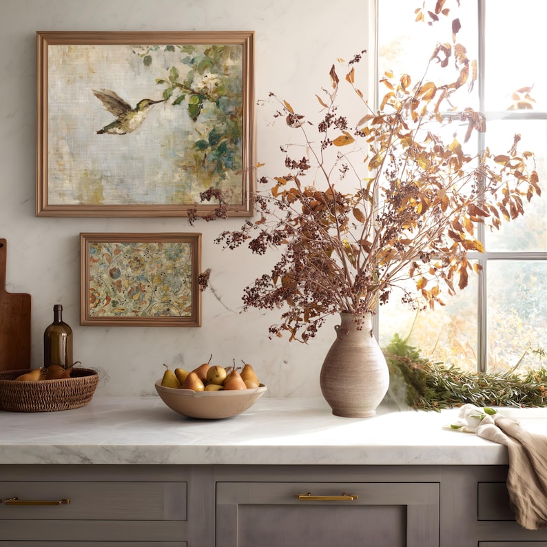 K&ouml;nnte beinhalten: Eine K&uuml;chenszene mit gerahmten Kunstwerken, einer Vase mit getrockneten Zweigen und einer Sch&uuml;ssel mit Birnen. Die Kunstwerke zeigen einen Kolibri und florale Muster. Die Arbeitsplatte ist aus Marmor, und die Schr&auml;nke sind grau mit goldenen Griffen.