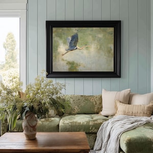 May include: A framed painting of a blue heron in flight, set against a muted green and beige background. The artwork is displayed above a green floral patterned sofa with decorative pillows and a wooden coffee table. The room has light blue paneling.