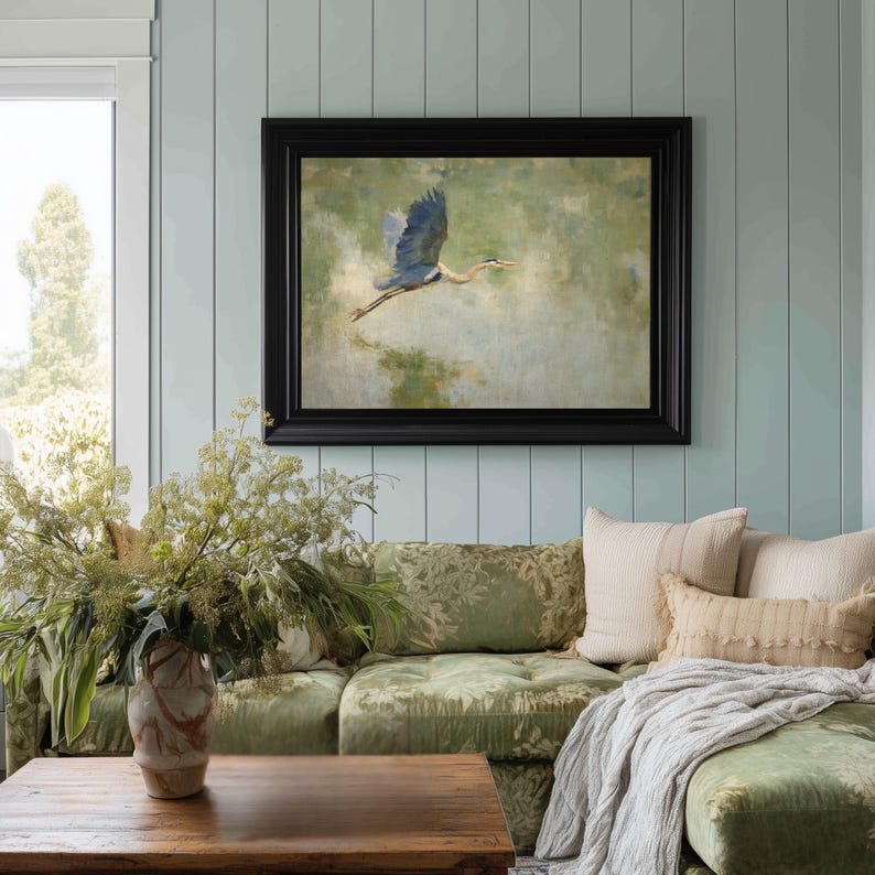 May include: A framed painting of a blue heron in flight, set against a muted green and beige background. The artwork is displayed above a green floral patterned sofa with decorative pillows and a wooden coffee table. The room has light blue paneling.