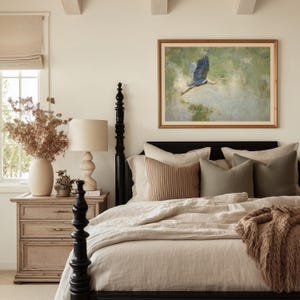 May include: A bedroom scene featuring a black four-poster bed with beige bedding and decorative pillows. A framed painting of a blue heron in flight hangs above the bed. A wooden nightstand with a lamp and vase of dried flowers completes the cozy, neutral-toned space.