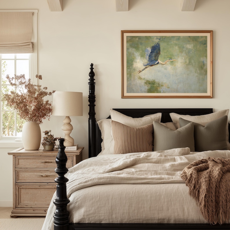 May include: A bedroom scene featuring a black four-poster bed with beige bedding and decorative pillows. A framed painting of a blue heron in flight hangs above the bed. A wooden nightstand with a lamp and vase of dried flowers completes the cozy, neutral-toned space.
