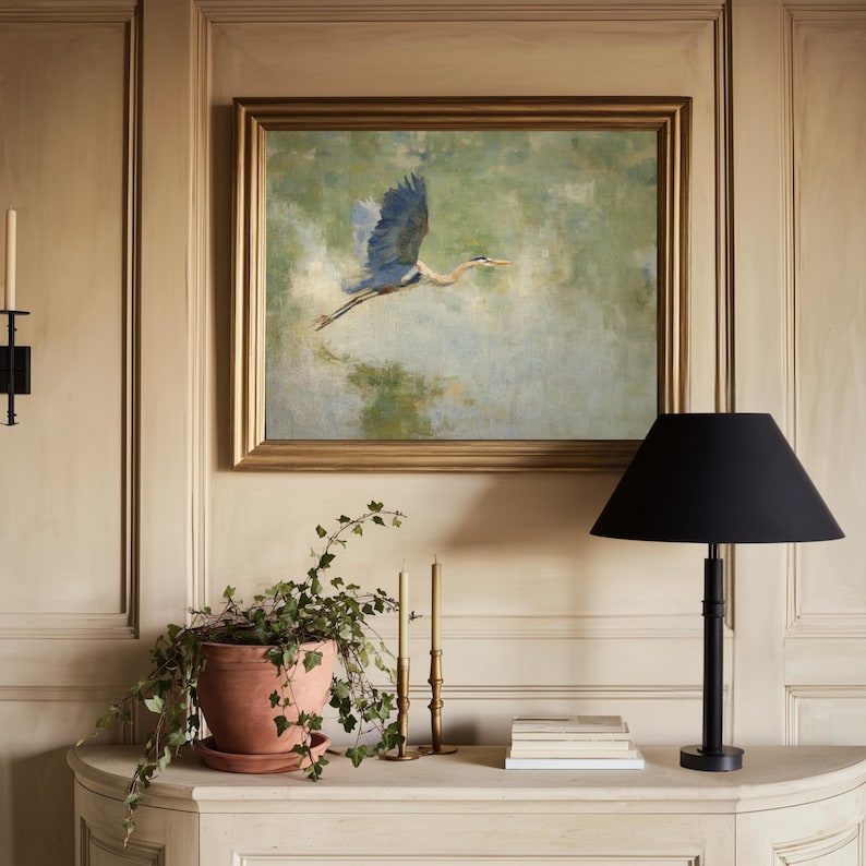 May include: An oil painting of a blue heron in flight hangs above a white console table. A black lamp with a conical shade sits on the table, along with a terracotta pot with greenery, candles, and books. The painting has a gold frame.