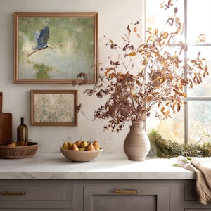 May include: A kitchen scene featuring a framed painting of a blue heron in flight, a smaller framed artwork, and a vase with dried branches. A bowl of pears sits on the countertop, alongside a woven basket and a bottle.