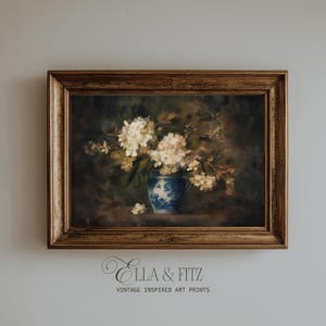 Pode incluir: Uma impressão de arte vintage emoldurada com uma natureza morta de flores brancas num vaso azul e branco. A obra de arte está sobre um fundo escuro e emoldurada numa moldura de madeira dourada. O texto "Ella & Fitz Vintage Inspired Art Prints" está na parte inferior.