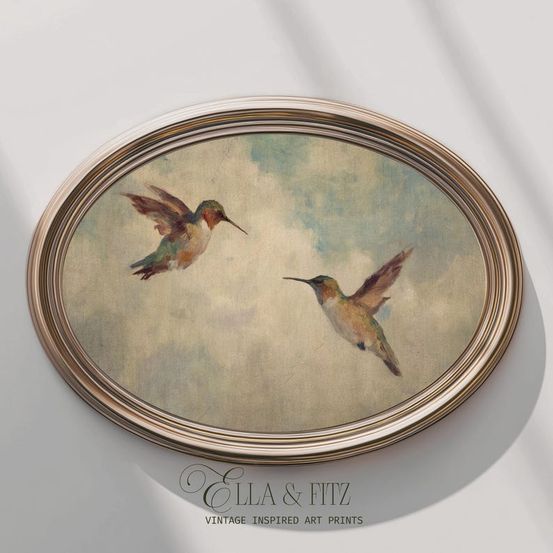 May include: An oval framed print depicting two hummingbirds in flight against a cloudy sky. The birds have brown, green, and red plumage. The frame is a light gold colour. The text "Ella & Fitz Vintage Inspired Art Prints" is at the bottom.