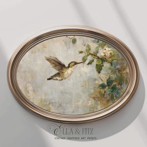 K&ouml;nnte beinhalten: Ein oval gerahmter Druck zeigt einen Kolibri im Flug, der sich einem bl&uuml;henden Zweig n&auml;hert. Das Kunstwerk hat einen weichen, malerischen Stil mit ged&auml;mpften Gr&uuml;nt&ouml;nen, Braunt&ouml;nen und Blaut&ouml;nen. Der Rahmen ist hellgoldfarben. Der Text "Ella & Fitz Vintage Inspired Art Prints" steht unten.