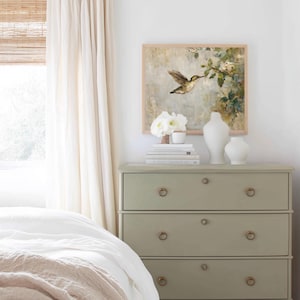 K&ouml;nnte beinhalten: Ein Schlafzimmer mit einer salbeigr&uuml;nen Kommode mit Messing-Ringgriffen, auf der B&uuml;cher, wei&szlig;e Vasen und ein gerahmtes Kolibri-Gem&auml;lde stehen. Weiche wei&szlig;e Vorh&auml;nge und ein Bett mit einer wei&szlig;en Bettdecke und einer beigen Decke vervollst&auml;ndigen die Szene.