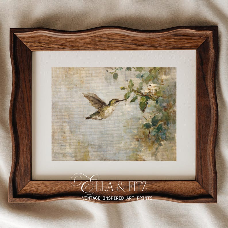 K&ouml;nnte beinhalten: Ein gerahmter Kunstdruck mit einem Kolibri im Flug, der nach einem bl&uuml;henden Zweig greift. Das Kunstwerk hat einen weichen, malerischen Stil mit ged&auml;mpften Blau-, Gr&uuml;n- und Wei&szlig;t&ouml;nen. Der Rahmen ist aus dunklem Holz mit dekorativem Rand. Der Text "Ella & Fitz" und "Vintage Inspired Art Prints" befindet sich unten.