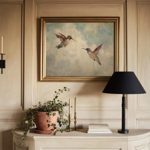 May include: A framed painting of two hummingbirds in flight against a cloudy sky hangs above a white console table. A black lamp, terracotta pot with greenery, candles, and books are on the table. The room has light-coloured panelling.