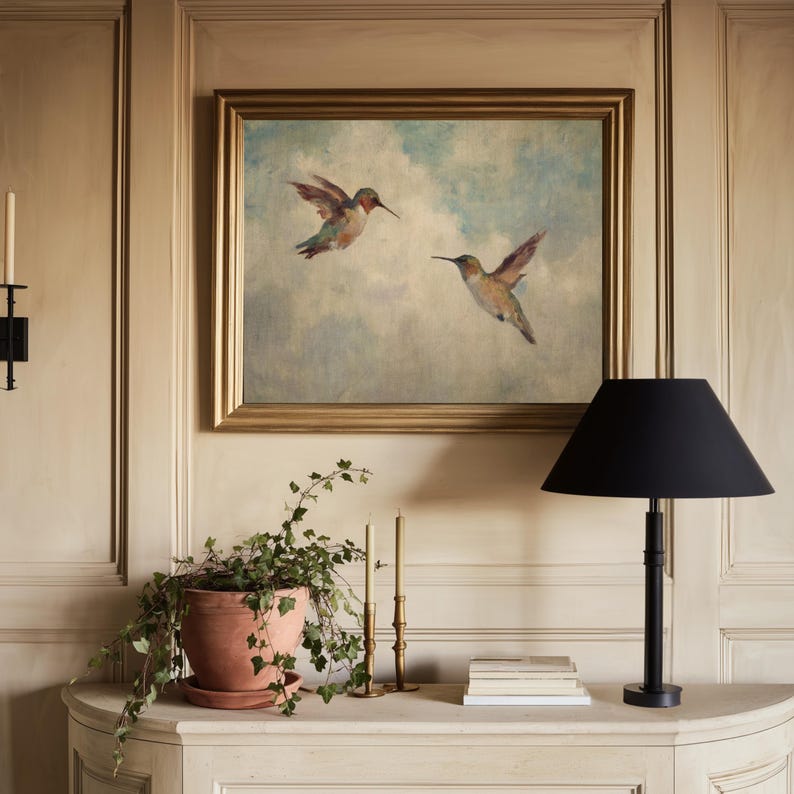 May include: A framed painting of two hummingbirds in flight against a cloudy sky hangs above a white console table. A black lamp, terracotta pot with greenery, candles, and books are on the table. The room has light-coloured panelling.