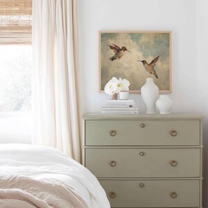 May include: A framed watercolour painting of two hummingbirds in flight hangs above a sage green chest of drawers. The chest has three drawers with brass ring pulls. White vases and books are on top. A bed with white linens is in the foreground.