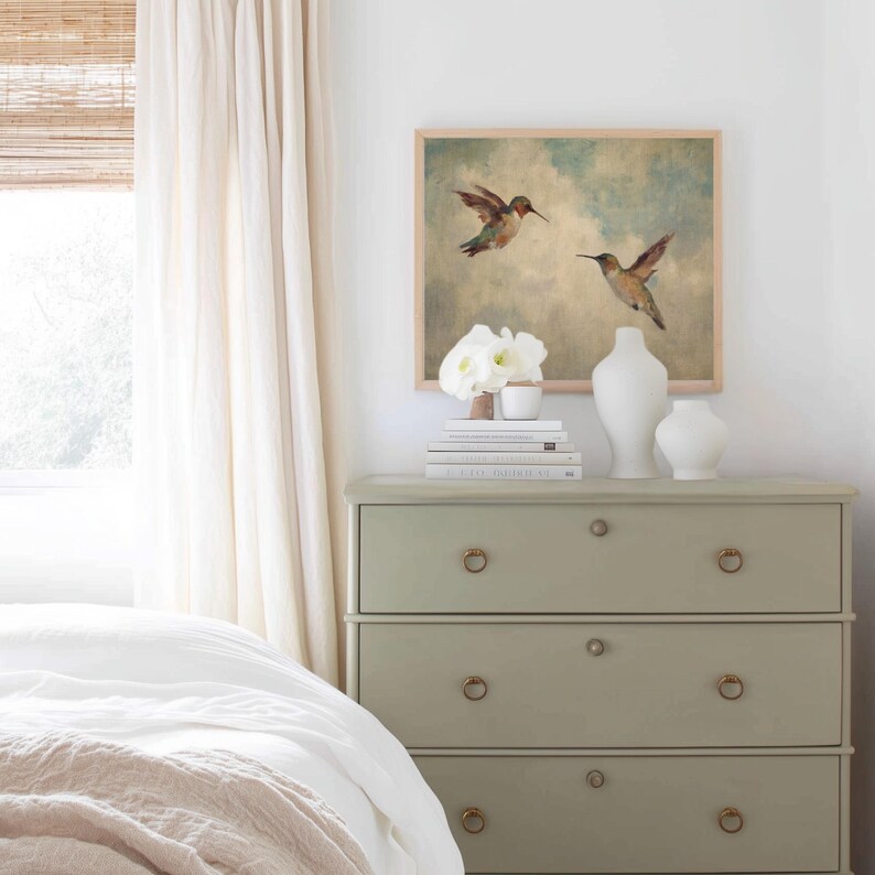 May include: A framed watercolour painting of two hummingbirds in flight hangs above a sage green chest of drawers. The chest has three drawers with brass ring pulls. White vases and books are on top. A bed with white linens is in the foreground.