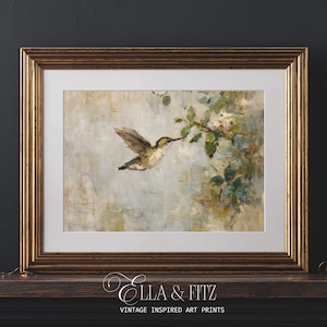 K&ouml;nnte beinhalten: Ein gerahmter Kunstdruck mit einem Kolibri im Flug, der sich einem bl&uuml;henden Zweig n&auml;hert. Das Kunstwerk hat einen weichen, malerischen Stil mit ged&auml;mpften T&ouml;nen von Gr&uuml;n, Braun und Blau. Der Rahmen ist goldfarben und der Druck tr&auml;gt die Aufschrift "Ella & Fitz Vintage Inspired Art Prints."