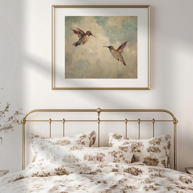 May include: A framed print depicting two hummingbirds in flight against a cloudy sky. The artwork is in a gold frame and hangs above a bed with floral bedding. The hummingbirds have green, red, and brown feathers.