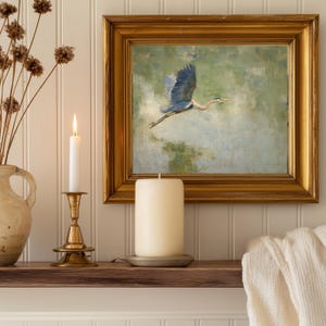 May include: A framed painting of a blue heron in flight hangs above a wooden shelf. A lit white candle sits in a brass candlestick, and a large pillar candle rests on a metal plate. Dried flowers are in a ceramic vase.