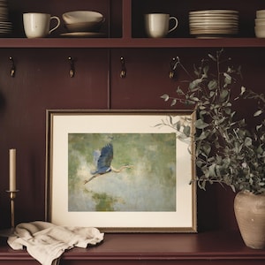 May include: A framed art print featuring a blue heron in flight against a soft green and blue background. The artwork is displayed in a gold-toned frame and rests on a dark red surface. A candle holder and a vase with greenery are also visible.
