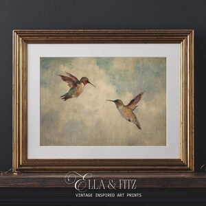 May include: A framed art print depicting two hummingbirds in flight against a cloudy sky. The birds have green, red, and brown feathers. The frame is gold-coloured, and the print is labelled "Ella & Fitz Vintage Inspired Art Prints."