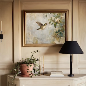 K&ouml;nnte beinhalten: Ein gerahmtes Gem&auml;lde eines Kolibris im Flug mit goldenem Rahmen h&auml;ngt &uuml;ber einem wei&szlig;en Schrank. Eine schwarze Lampe mit konischem Schirm steht auf dem Schrank, zusammen mit einer Topfpflanze, Kerzen und B&uuml;chern.