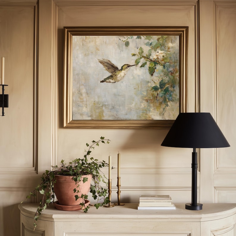 K&ouml;nnte beinhalten: Ein gerahmtes Gem&auml;lde eines Kolibris im Flug mit goldenem Rahmen h&auml;ngt &uuml;ber einem wei&szlig;en Schrank. Eine schwarze Lampe mit konischem Schirm steht auf dem Schrank, zusammen mit einer Topfpflanze, Kerzen und B&uuml;chern.