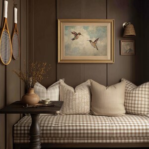 May include: A warm interior with a bench upholstered in a brown and white checked fabric. Cushions in neutral shades are arranged on the bench. A framed picture of hummingbirds is displayed, and two vintage tennis rackets hang on the wall.